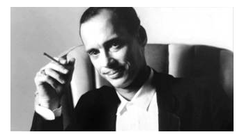 debonaire John Waters sitting on a chair with a cigarette in his hand, raised in the air near head level